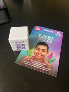 Cubo branco com código QR do site 'raras.pt' junto a um folheto promocional colorido datado de 28 FEV 2025. O folheto mostra uma pessoa sorridente com as mãos pintadas em várias cores, tendo ao fundo um gradiente azul e rosa. No topo do folheto está escrito 'MUITO MAIS DO QUE PODE IMAGINAR', numa publicação da Raras Portugal.