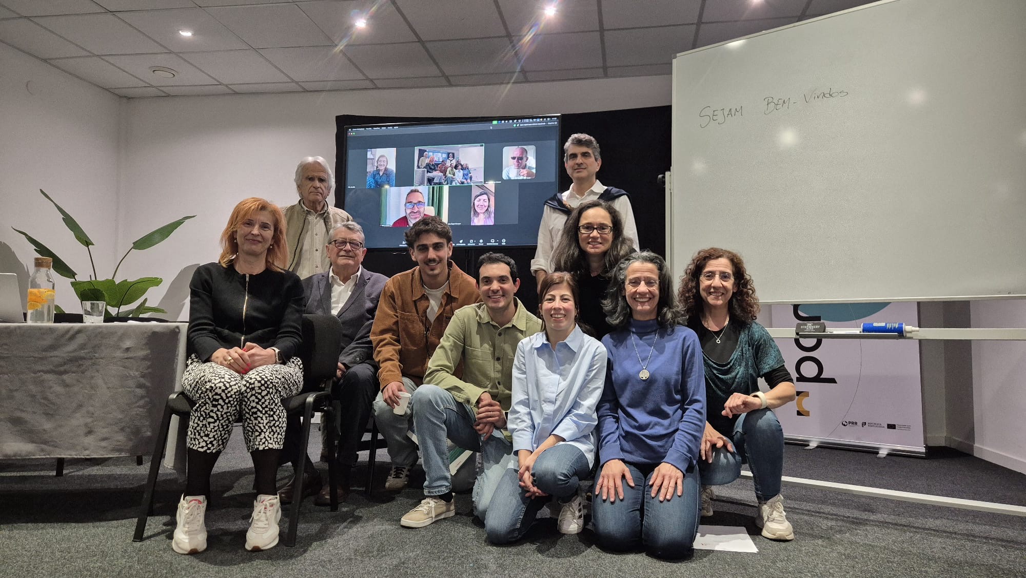 Fotografia da Assembleia Geral da APDIP 2025. Na sala de reunião, oito pessoas estão presentes fisicamente, posando para a foto em grupo, com algumas pessoas sentadas e outras em pé. Ao fundo, uma tela grande mostra participantes conectados remotamente por videoconferência.