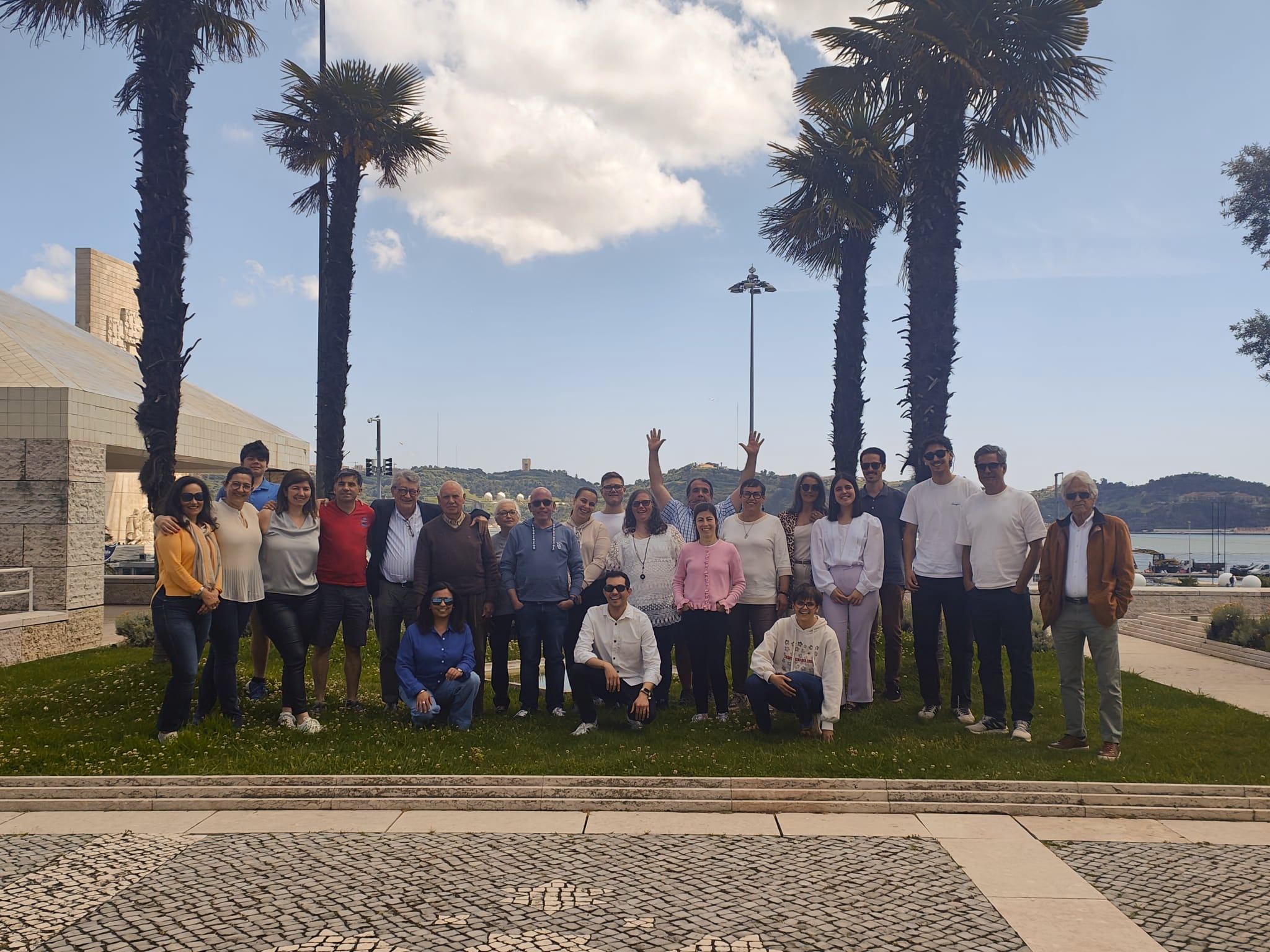 Fotografia de grupo de aproximadamente 20 pessoas ao ar livre numa área relvada com palmeiras, edifícios modernos e um fundo cénico com colinas e céu.