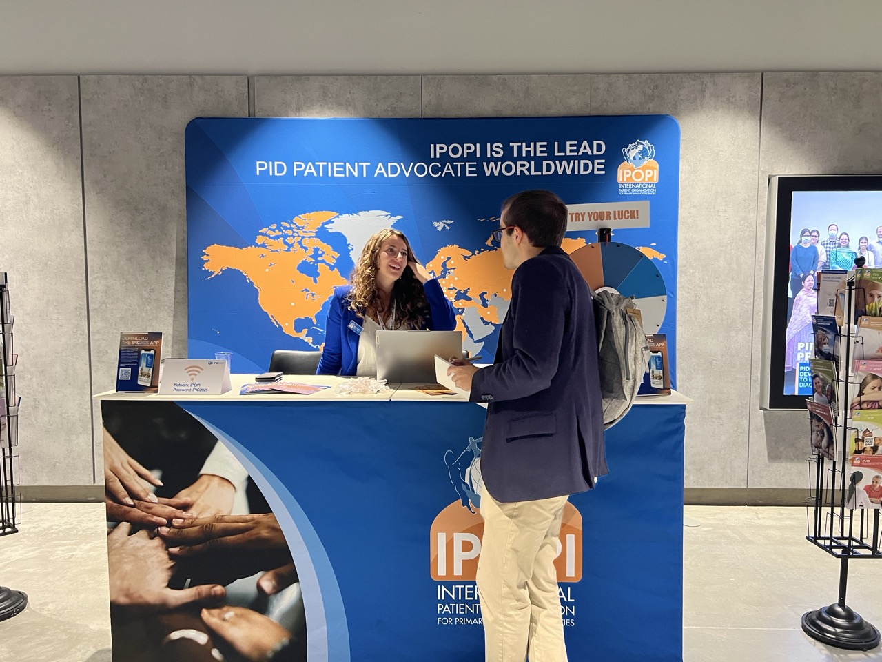 IPOPI booth at IPIC 2025 conference in Prague with staff member assisting visitor at information desk, featuring blue backdrop with world map and IPOPI branding as PID patient advocate worldwide.