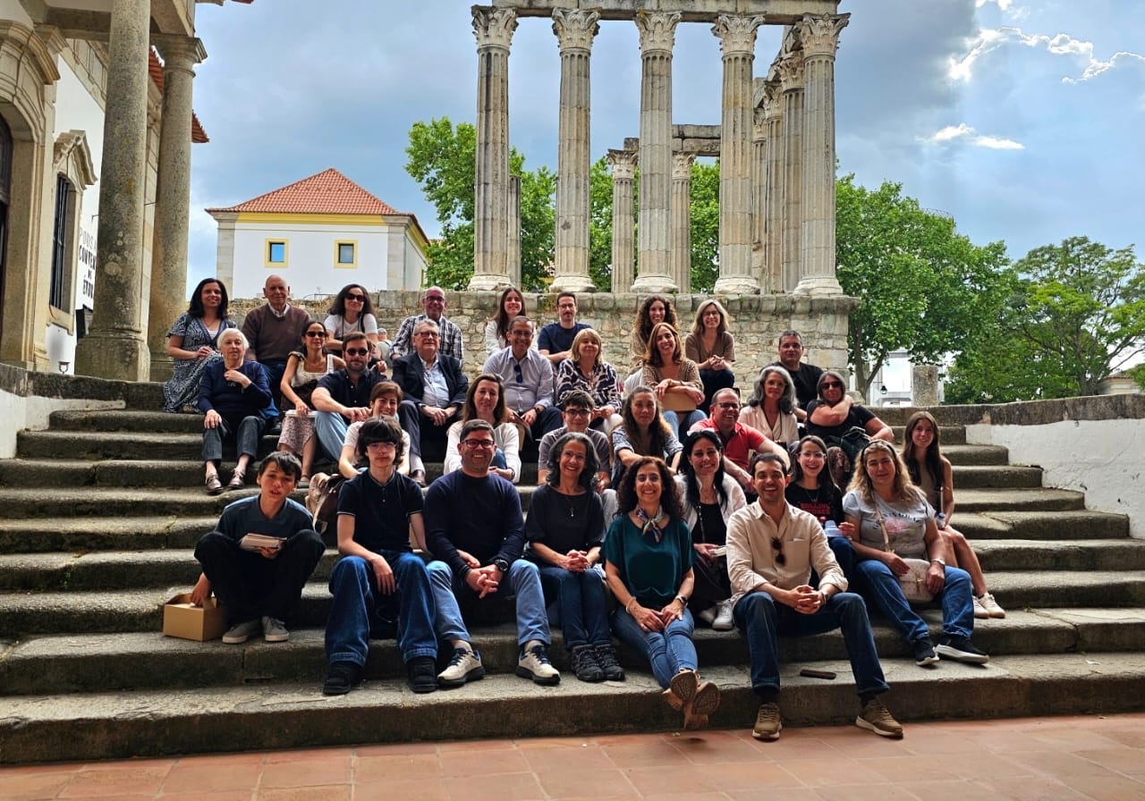 Fotografia de grupo de diversas pessoas sentadas e em pé nos degraus de pedra de um antigo templo romano, sob um céu parcialmente nublado e árvores verdes ao fundo.